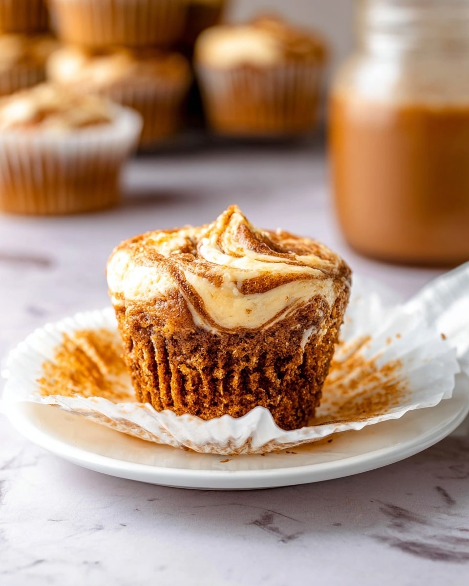 A single swirled muffin sits on a white plate with its white baking paper partly peeled down. The muffin has two visible layers: a textured brown base that looks moist and crumbly, topped by a creamy, light beige swirl layer with a soft, smooth texture. In the blurred background, there are more muffins and a glass jar with a brown drink on a white marbled surface. The light is soft and natural, highlighting the muffin’s swirl pattern and moist crumb, photo taken with an iphone --ar 4:5 --v 7