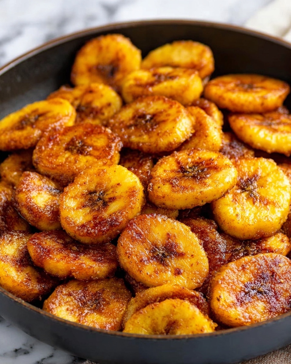 The image shows a close-up view of many small, round fried plantain slices with a rich golden brown color and a slightly crispy texture. Each piece has some areas darker than others, indicating a perfect caramelized surface with a light sprinkle of spices or seasoning on top. The plantains are softly piled together, filling a pan that has a dark interior but is resting on a white marbled surface. Light shines gently on the plantains, highlighting their shiny, cooked surface. Photo taken with an iphone --ar 4:5 --v 7