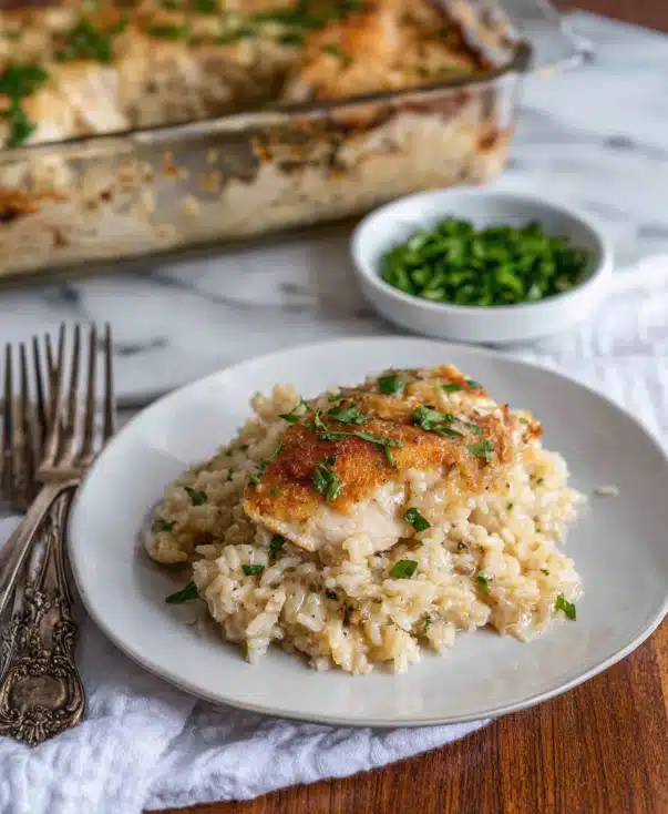 A white plate holds a serving of creamy rice with a piece of light brown cooked chicken on top. The chicken is smooth and slightly shiny, with some black pepper and small green herbs sprinkled over it. The rice looks soft and moist with a few visible grains and bits of seasoning mixed in. The food rests on a white cloth napkin with two silver forks beside the plate. The background is blurred but shows a brown wooden surface and a glass baking dish filled with more rice and chicken. photo taken with an iphone --ar 4:5 --v 7