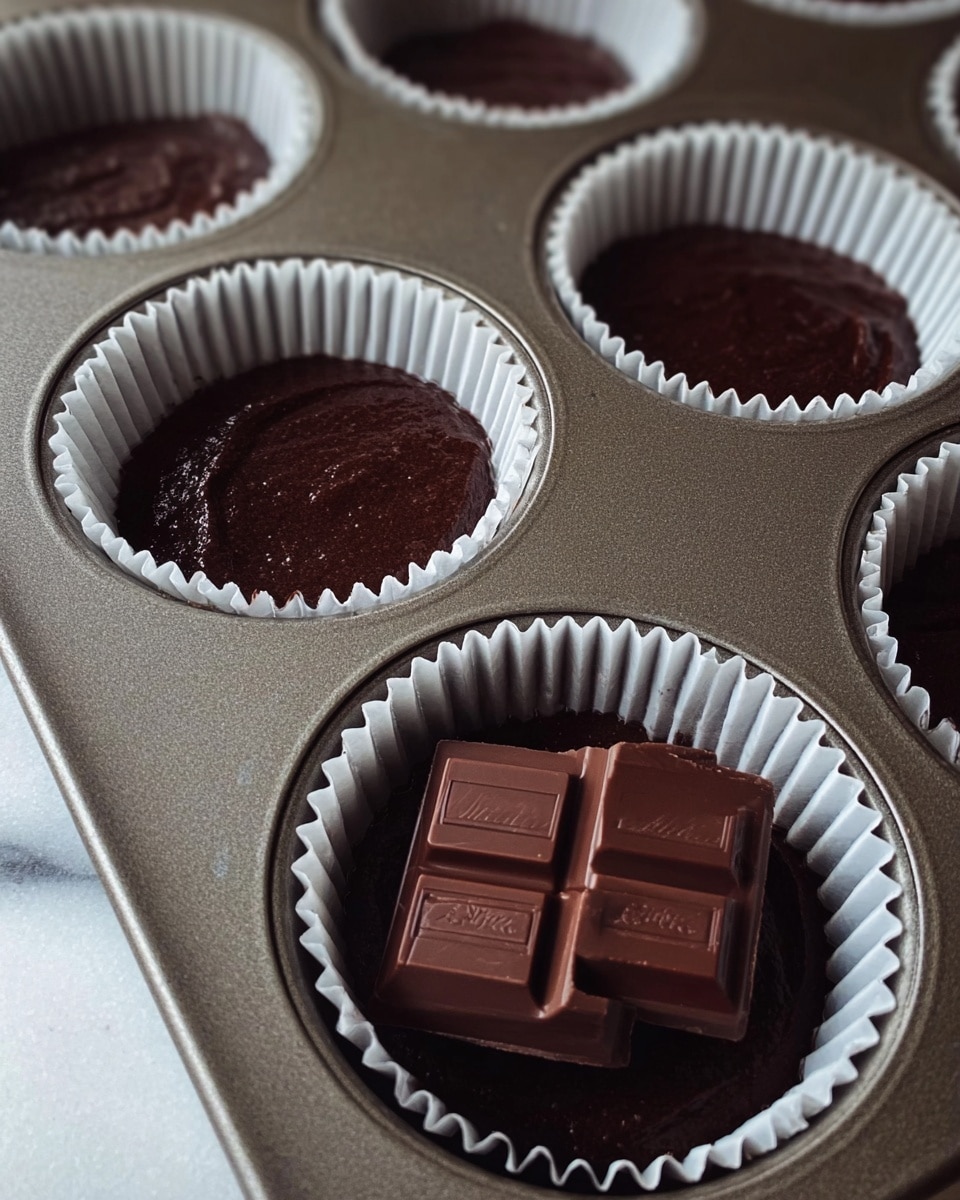 The image shows a close-up of a baking tray with six round white paper liners filled with smooth, dark brown chocolate batter. One liner in the foreground holds two pieces of solid dark chocolate placed on top of the batter. The chocolate pieces are shiny and rectangular with some text visible on them. The tray rests on a white marbled surface with soft natural lighting. The overall look is clean and focused on the rich chocolate texture. photo taken with an iphone --ar 4:5 --v 7