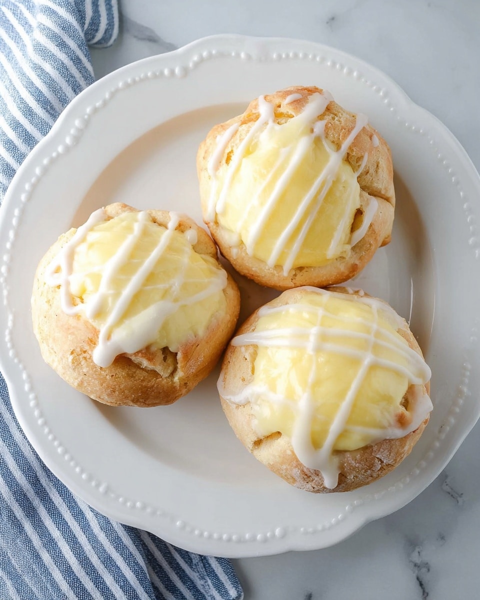 The image shows three round pastry buns on a white plate with a scalloped edge, placed on a white marbled surface. Each bun has a light golden-brown crust with a slightly rough texture, forming the bottom and sides. On top of each bun is a thick, creamy pale yellow filling with a smooth, glossy surface. White icing is drizzled in thin, uneven lines over the filling and the crust, adding a shiny contrast. The buns are arranged with two close at the bottom right and one slightly higher at the top left. A glimpse of a blue-striped fabric is visible at the bottom left corner. Photo taken with an iphone --ar 4:5 --v 7