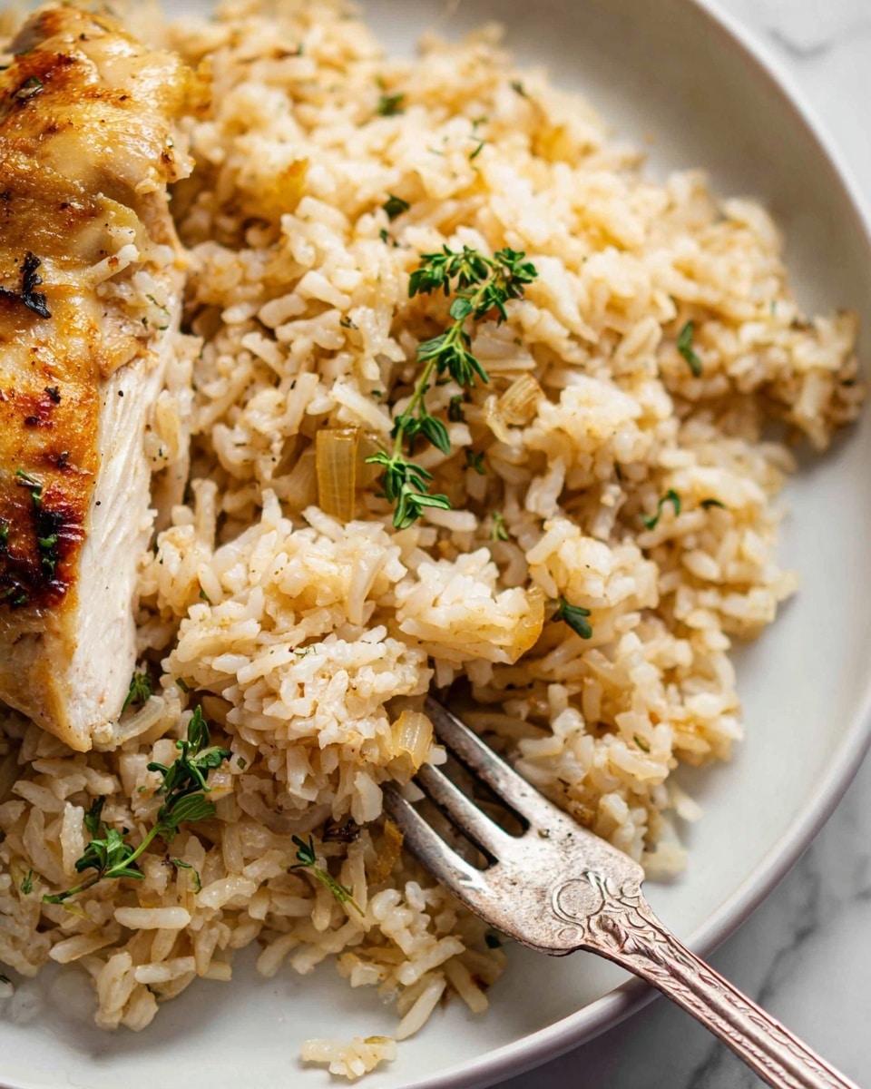 The image shows a close-up of a white plate with two main layers. The bottom layer is a bed of fluffy light brown rice mixed with small pieces of cooked onion and a few sprigs of fresh green thyme scattered on top. The second layer partially visible on the left side is a piece of golden-brown cooked chicken with a slightly crispy skin. A silver fork with an ornate handle is placed on the right side, pushing into the rice, with its tip under the chicken slice. The setting includes a white marbled surface under the plate. Photo taken with an iphone --ar 4:5 --v 7