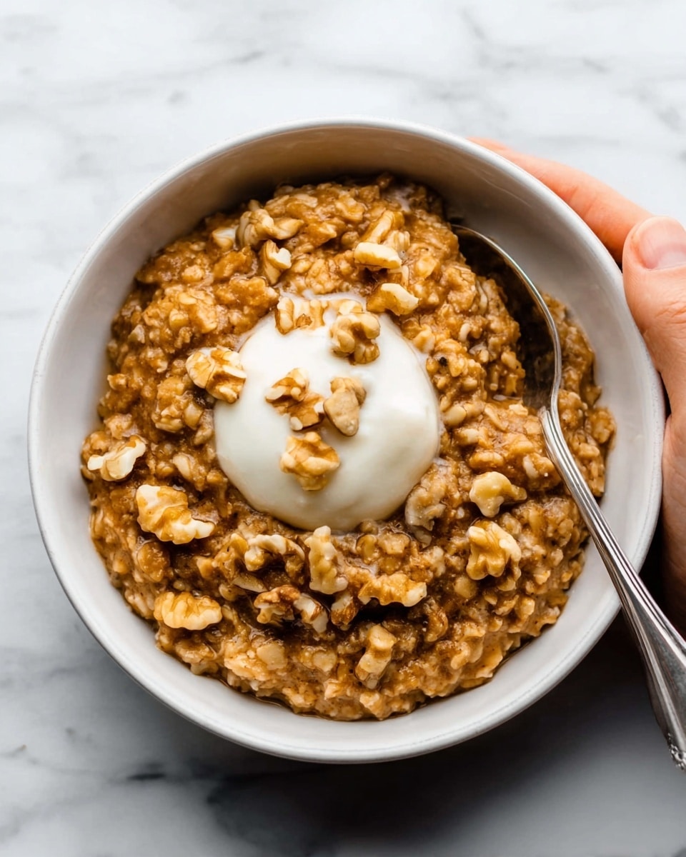 A white bowl filled with thick, brown oatmeal that has visible pieces of walnuts mixed throughout, creating a rough and chunky texture on top. To one side of the oatmeal, there is a smooth dollop of pale cream or yogurt sitting on the surface. The bowl is placed on a white marbled texture with a silver spoon resting nearby. A woman's hand is gently holding the bowl from one edge. Photo taken with an iphone --ar 4:5 --v 7