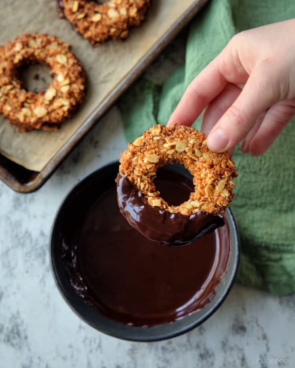 A woman's hand holding a round cookie with a hole in the middle, partially dipped in shiny dark chocolate. The cookie has a rough texture with nuts or oats on the surface, giving it a crunchy look. Below, there is a black bowl filled with smooth dark chocolate. Next to the bowl, on a metal baking tray lined with baking paper, two similar cookies rest, showing their golden-brown color and nutty texture. A green cloth is placed behind the bowl on a white marbled surface. Photo taken with an iphone --ar 4:5 --v 7
