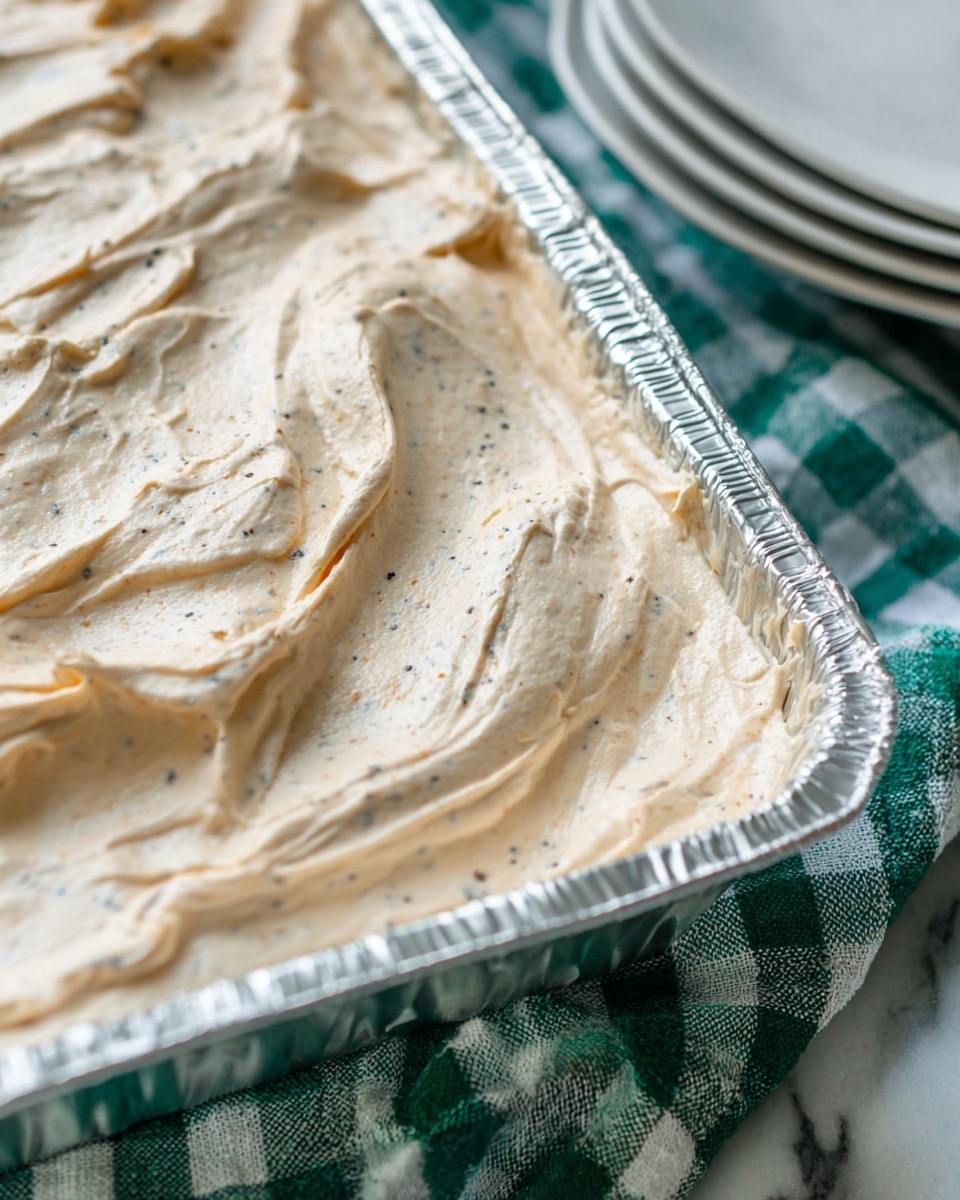 A close-up view of a thick, creamy, light beige spread with specks of darker seasoning swirled smoothly inside a shiny silver foil tray. The tray sits on a white marbled surface partially covered by a green and white checkered cloth. To the right edge, there is a stack of plain white plates slightly out of focus. The texture of the spread looks soft and airy with some gentle peaks and folds photo taken with an iphone --ar 4:5 --v 7