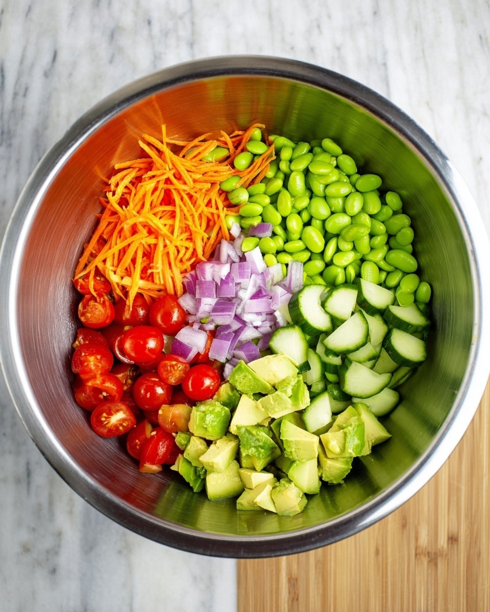 A large silver mixing bowl sits on a white marbled surface. Inside, the vegetables are separated into six sections: bright green edamame beans cover almost half the bowl to the right, thin orange carrot strips sit above them, small light purple chopped onions are in the center, fresh dark green cucumber slices are to the left, juicy red cherry tomato halves below them, and chunky pale green avocado pieces are above the cucumber slices. The colors and textures are fresh and vibrant, ready to be mixed. Photo taken with an iphone --ar 4:5 --v 7