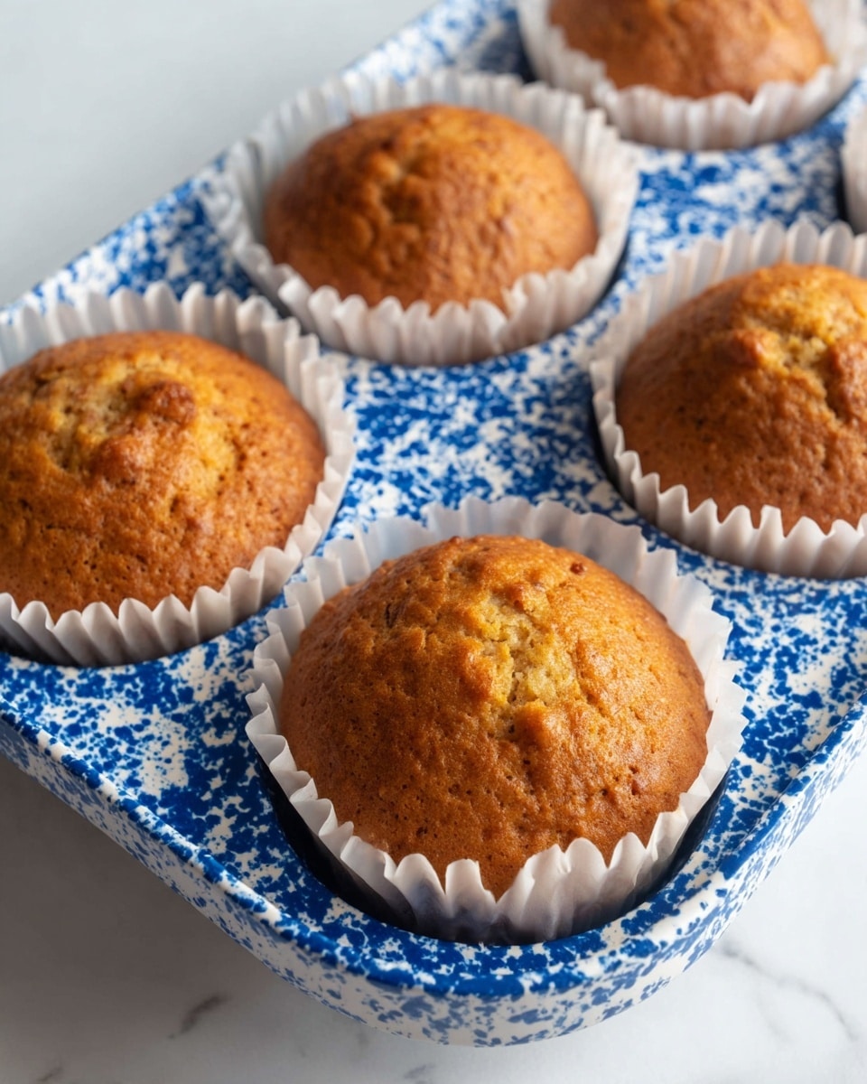 Four golden brown muffins with slightly rough tops and small cracks on the surface sit inside a white cupcake pan with blue speckled patterns. Each muffin is nestled in white paper liners, filling each round cup of the pan. The background is a white marbled surface. photo taken with an iphone --ar 4:5 --v 7