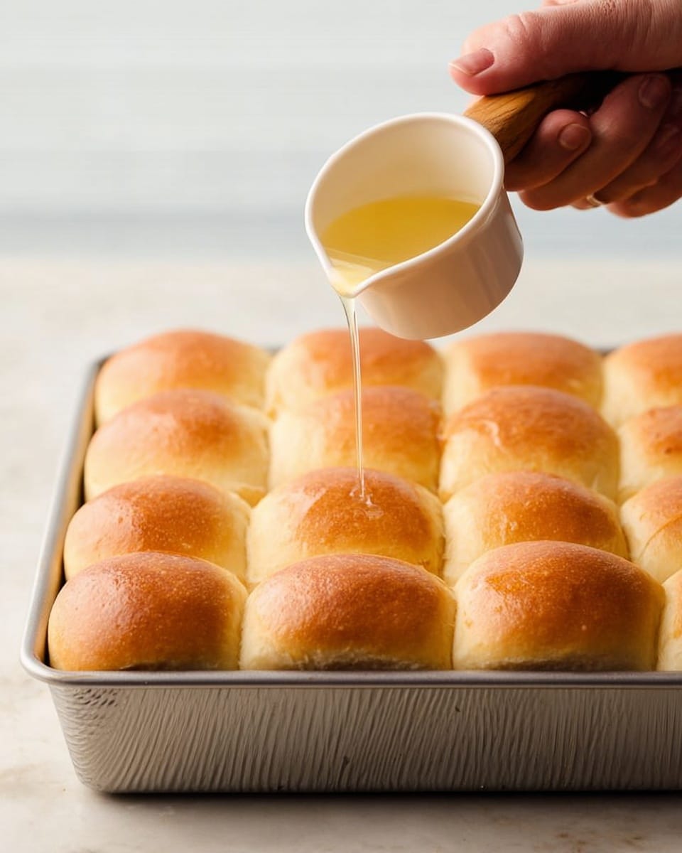 The image shows a close-up view of 16 soft golden-brown dinner rolls arranged in 4 rows and 4 columns inside a metal baking pan with slightly textured sides. A woman's hand holds a small white cup with a wooden handle above the rolls, pouring a shiny, light-yellow liquid evenly over them. The rolls have smooth, slightly rounded tops with a light crust. The background and surface are white with a marbled texture, providing a clean and bright setting. Photo taken with an iphone --ar 4:5 --v 7