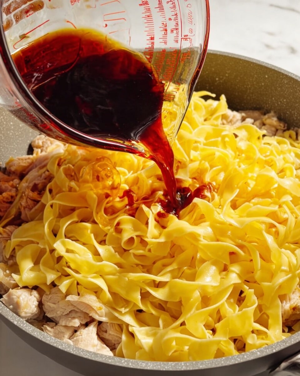 A gray cooking pan contains a layer of cooked light yellow flat pasta ribbons, resting on top of a layer of light beige cooked meat pieces. A clear glass measuring cup is tilted above the pan, pouring a thick dark reddish-brown sauce over the pasta. The pan is placed on a white marbled surface. photo taken with an iphone --ar 4:5 --v 7