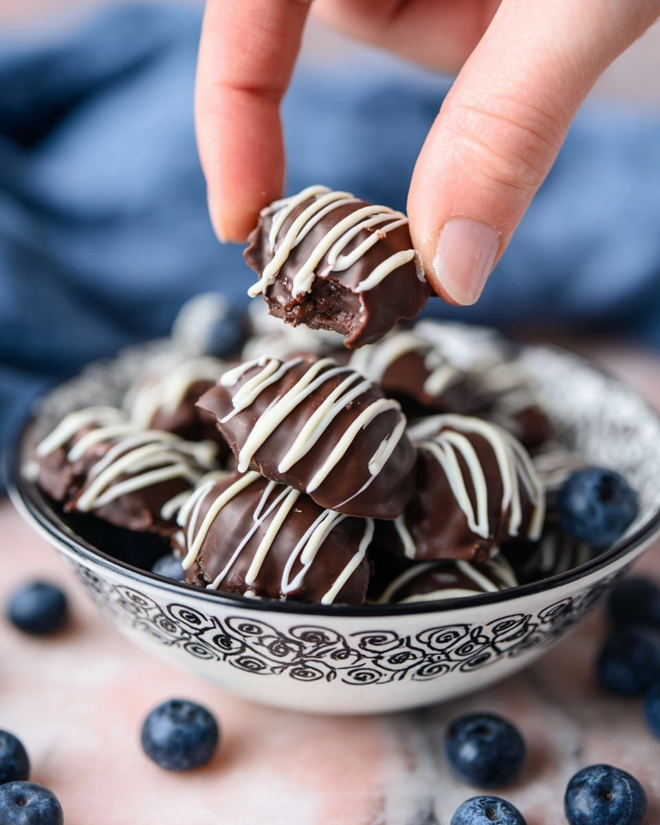 A close-up image shows a woman's hand holding a dark brown, chocolate-covered cluster with a smooth texture, drizzled with thin, white chocolate lines across the top. Below, a pile of similar chocolate clusters sits inside a white bowl with a decorative black pattern on the rim. Around the bowl, there are many fresh, plump blueberries with a deep blue color, scattered on a white marbled surface. The background is softly blurred, drawing attention to the detailed textures of the chocolate and berries. photo taken with an iphone --ar 4:5 --v 7