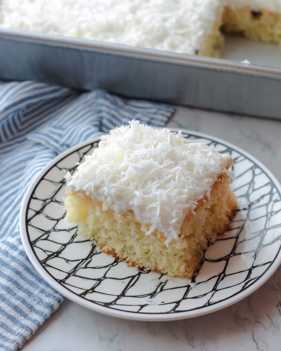 A square piece of cake sits on a white plate with a black diamond pattern along the edge. The cake has two layers: the bottom layer is a light yellow sponge cake with a soft, crumbly texture, and the top layer is a thick, fluffy white frosting sprinkled with shredded coconut, giving it a slightly rough texture. In the background, there is a metal baking pan containing the rest of the cake with the same white frosting layer visible, all placed on a white marbled surface next to a blue and white striped cloth. Photo taken with an iphone --ar 4:5 --v 7