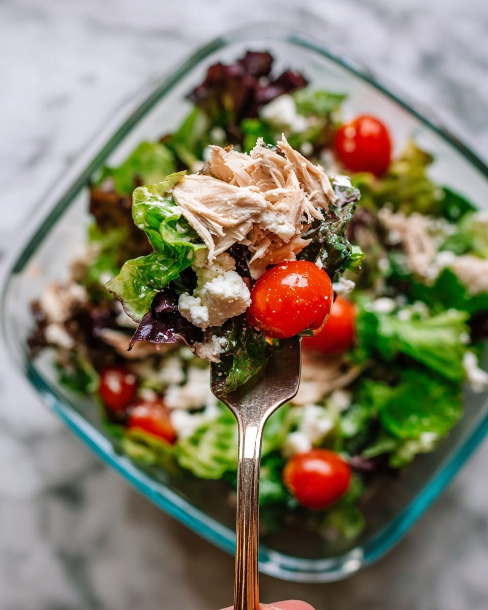 A fork holds a fresh salad with four main layers: the bottom layer has mixed green and purple lettuce leaves with a soft texture, above it there are bright red cherry tomatoes, then light beige shredded cooked chicken pieces with a slightly fibrous texture, and on top, dollops of white creamy cheese. The salad is in a clear glass dish sitting on a white marbled surface, and a woman's hand is holding the fork that is centered in the image, with the salad background softly out of focus. photo taken with an iphone --ar 4:5 --v 7