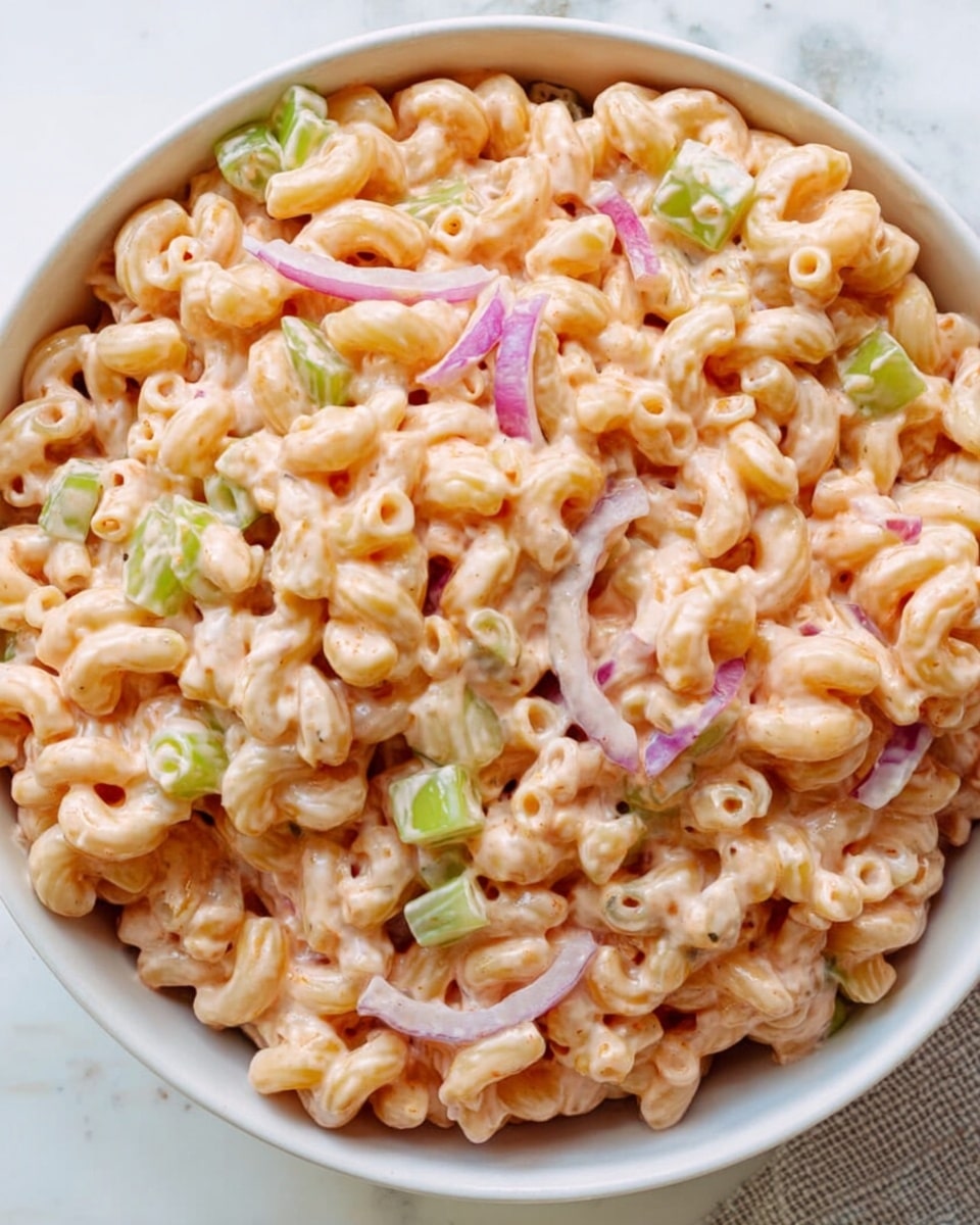 A close-up image showing a spoon lifting a portion of creamy pasta salad from a white bowl, the pasta shapes are curved macaroni coated with a smooth, orange-colored sauce; visible in the mix are small pieces of light green celery and small chunks of purple-red onion, all sitting on a white marbled surface. photo taken with an iphone --ar 4:5 --v 7