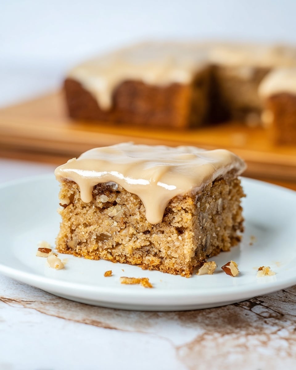 The image shows a single square piece of light brown cake with visible nut pieces inside, placed on a white plate. The cake has one layer with a smooth layer of creamy, light beige frosting on top that drips slightly down the sides. There are crumbs scattered on the plate around the piece. In the background, a larger section of the same cake with the same frosting is slightly out of focus. The plate sits on a wooden surface with a white marbled texture visible underneath. Photo taken with an iphone --ar 4:5 --v 7