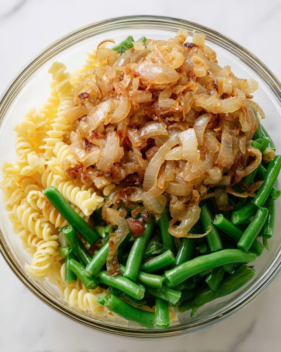 The image shows a clear glass bowl filled with three main layers of food. The bottom layer is a bed of spiral pasta, creamy yellow in color and textured with small ridges. The middle layer consists of cooked green beans, chopped into small, even pieces, showing a bright green color and fresh texture. The top layer is a generous pile of caramelized onions, light golden brown with some darker edges, soft and slightly translucent, covering part of the pasta and green beans. The bowl is placed on a white marbled surface. photo taken with an iphone --ar 4:5 --v 7