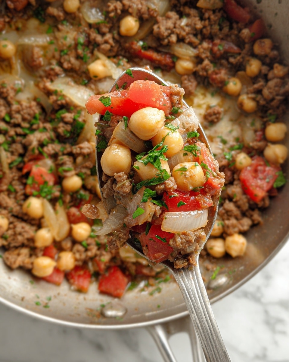 A close-up view of a metal pan filled with a mix of cooked ground meat, translucent cooked onion pieces, light beige round chickpeas, and small chunks of red tomatoes, all sprinkled with bright green chopped herbs. A silver spoon holds a portion of this mixture in the center, showing the same layers and colors of meat, onions, chickpeas, tomatoes, and herbs clearly. The background is a white marbled texture. photo taken with an iphone --ar 4:5 --v 7
