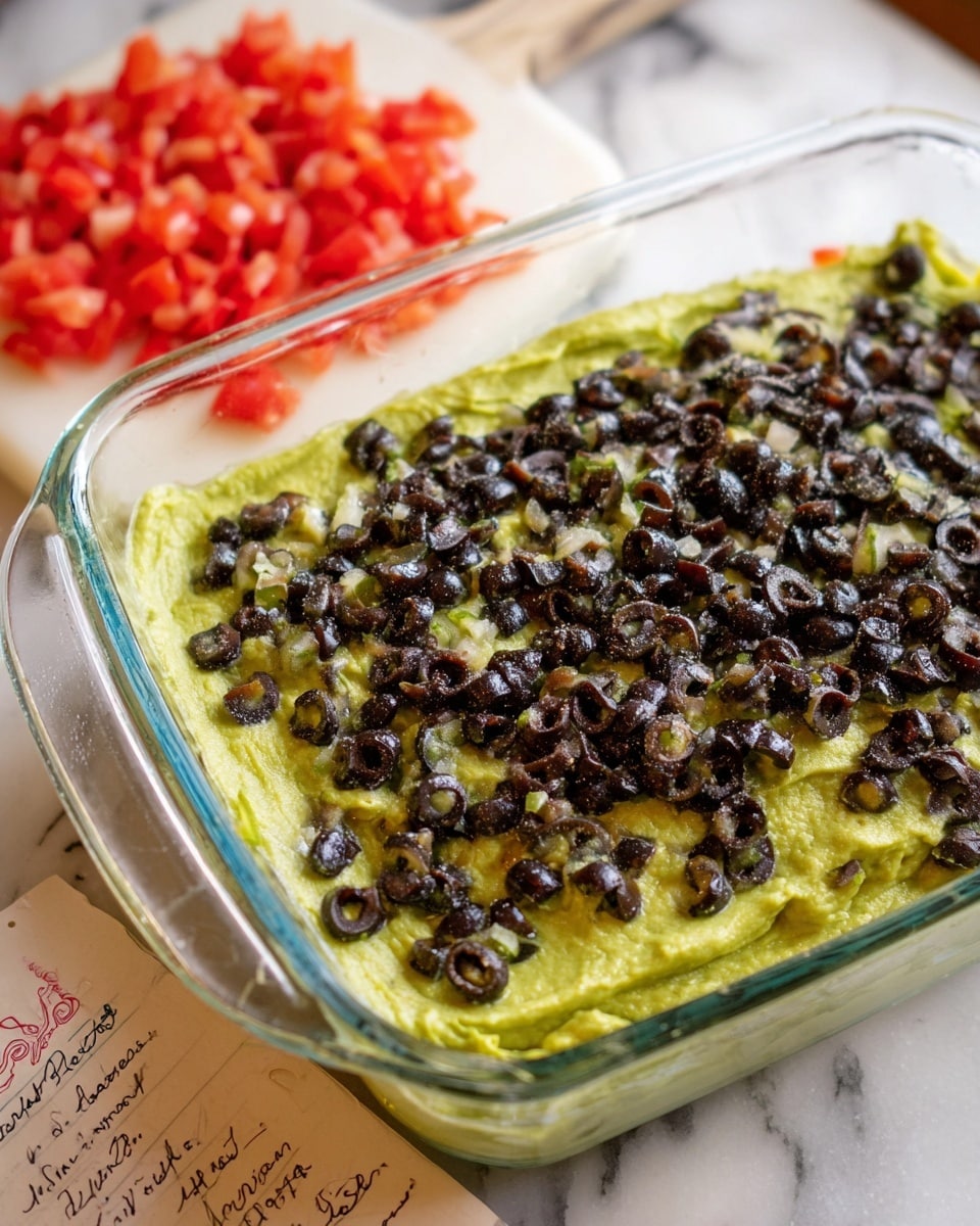 A clear glass baking dish filled with two visible layers; the bottom layer is smooth, bright green, likely mashed avocado or guacamole, spread evenly, and the top layer is a generous sprinkling of small, chopped black olives scattered thickly across the green base. The dish sits on a white marbled surface next to a white cutting board with finely diced red tomatoes. Part of a handwritten recipe card is visible beside the dish. Photo taken with an iphone --ar 4:5 --v 7
