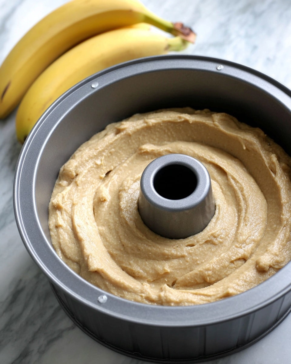 A gray bundt pan filled with light brown, smooth cake batter spread evenly inside. The batter's surface shows soft swirls making a slightly textured look. In the background, there are three yellow bananas resting on a white marbled surface. Photo taken with an iphone --ar 4:5 --v 7