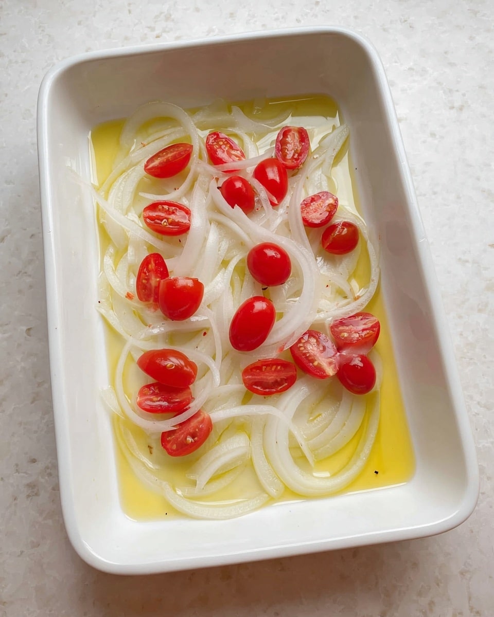 A white rectangular dish with light yellow oil spread evenly at the bottom, topped with thin, long white onion slices arranged loosely across the surface, and halved bright red cherry tomatoes scattered on top, creating a simple, fresh mix of colors and textures on a white marbled background photo taken with an iphone --ar 4:5 --v 7