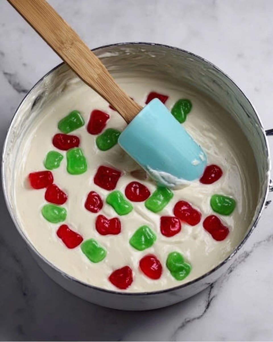 A white metal saucepan filled with a creamy white batter that looks smooth and thick. Scattered on top are many red and green jelly candies that are shiny and bright. A light blue silicone spatula with a wooden handle is resting inside the saucepan, partially covered in batter. The saucepan sits on a white marbled surface. photo taken with an iphone --ar 4:5 --v 7