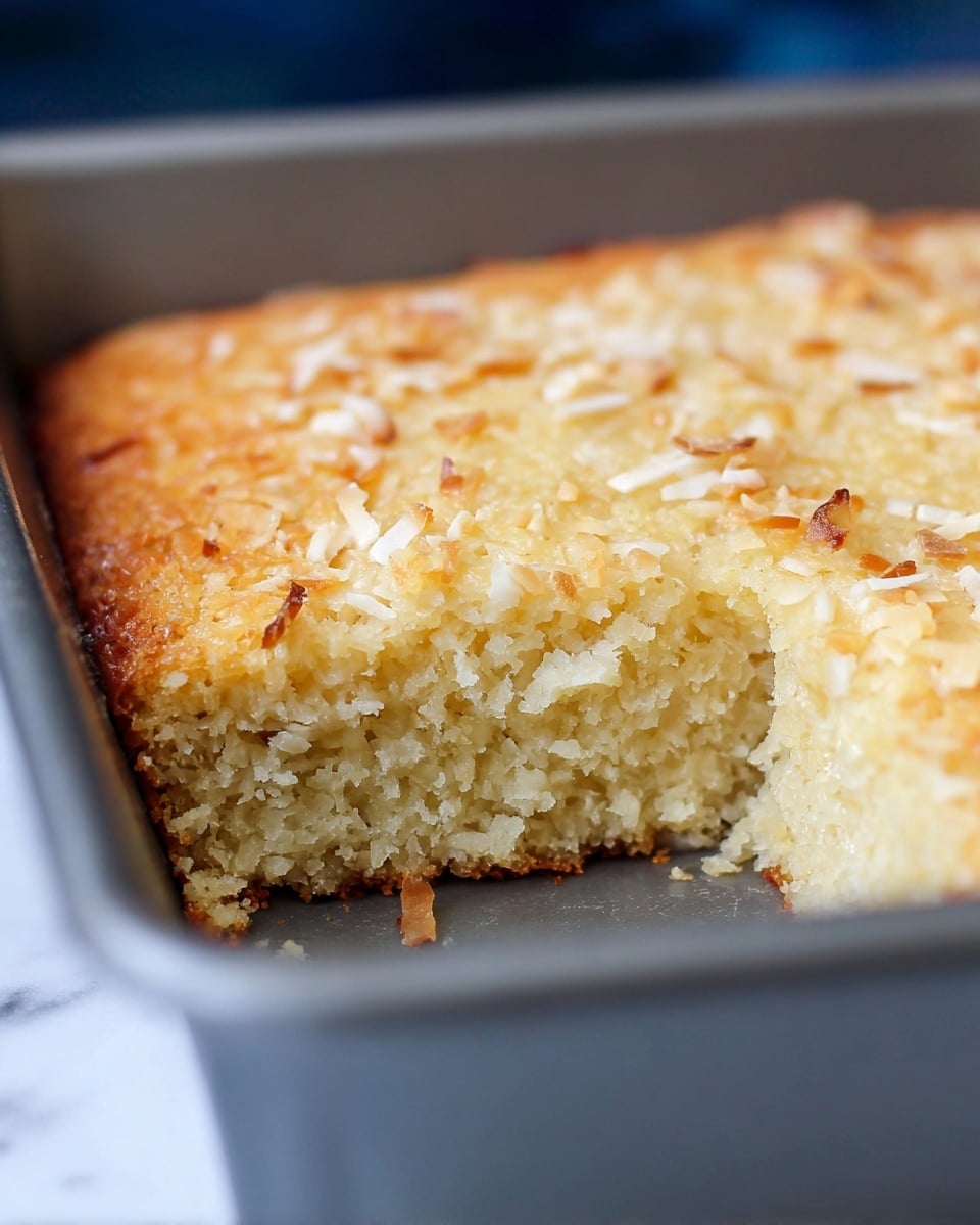 The image shows a close-up of one square piece removed from a thick, pale golden baked cake in a gray baking pan. The cake has a rough top textured with small toasted coconut flakes, showing a soft and crumbly inside with bits of coconut visible. The pan and the cake fill most of the frame, sitting on a white marbled surface with a blurry dark blue background visible at the top. Photo taken with an iphone --ar 4:5 --v 7