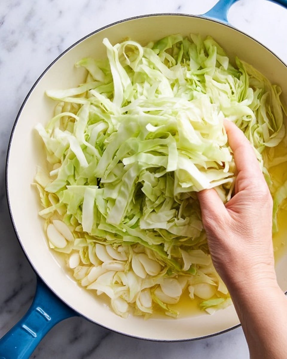 The image shows a white pan with a blue handle on a white marbled surface. Inside the pan, there are three layers: the bottom layer contains thin slices of garlic with a light yellow color in cooking oil, the middle layer has long ribbons of light green cabbage, and a woman's hand is adding more shredded cabbage from the top left corner into the pan. The cabbage has a crisp, slightly curled texture and pale green color, contrasting with the smooth garlic slices below. The light reflects softly on the shredded cabbage and garlic, making the ingredients look fresh and ready to cook. photo taken with an iphone --ar 4:5 --v 7