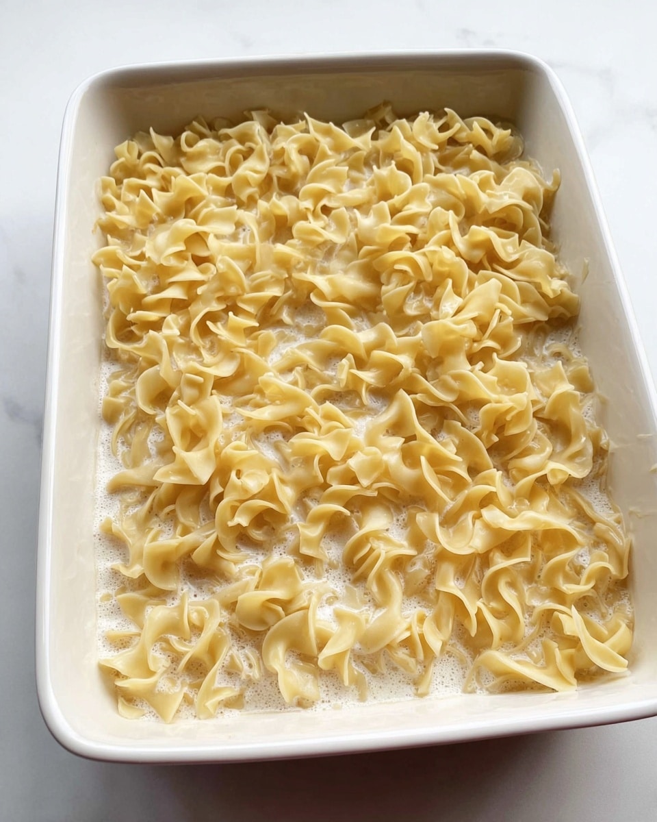 The image shows a white rectangular baking dish filled with cooked egg noodles and a creamy sauce. The noodles are yellowish and curly, spread evenly across the dish in one thick layer. The white sauce is visible between the noodles, with a slightly foamy texture that coats the pasta. The dish is placed on a white marbled surface. Photo taken with an iphone --ar 4:5 --v 7