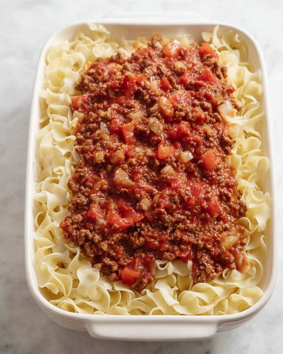 A white rectangular baking dish holds two visible layers, with the bottom layer consisting of light yellow cooked egg noodles that have a soft and curled texture covering the dish's base. The top layer features a chunky meat sauce with visible pieces of ground beef, diced tomatoes, and small bits of onion, creating a reddish-brown and red mix that covers the noodles in the center and extends slightly outward. The dish rests on a white marbled surface. photo taken with an iphone --ar 4:5 --v 7