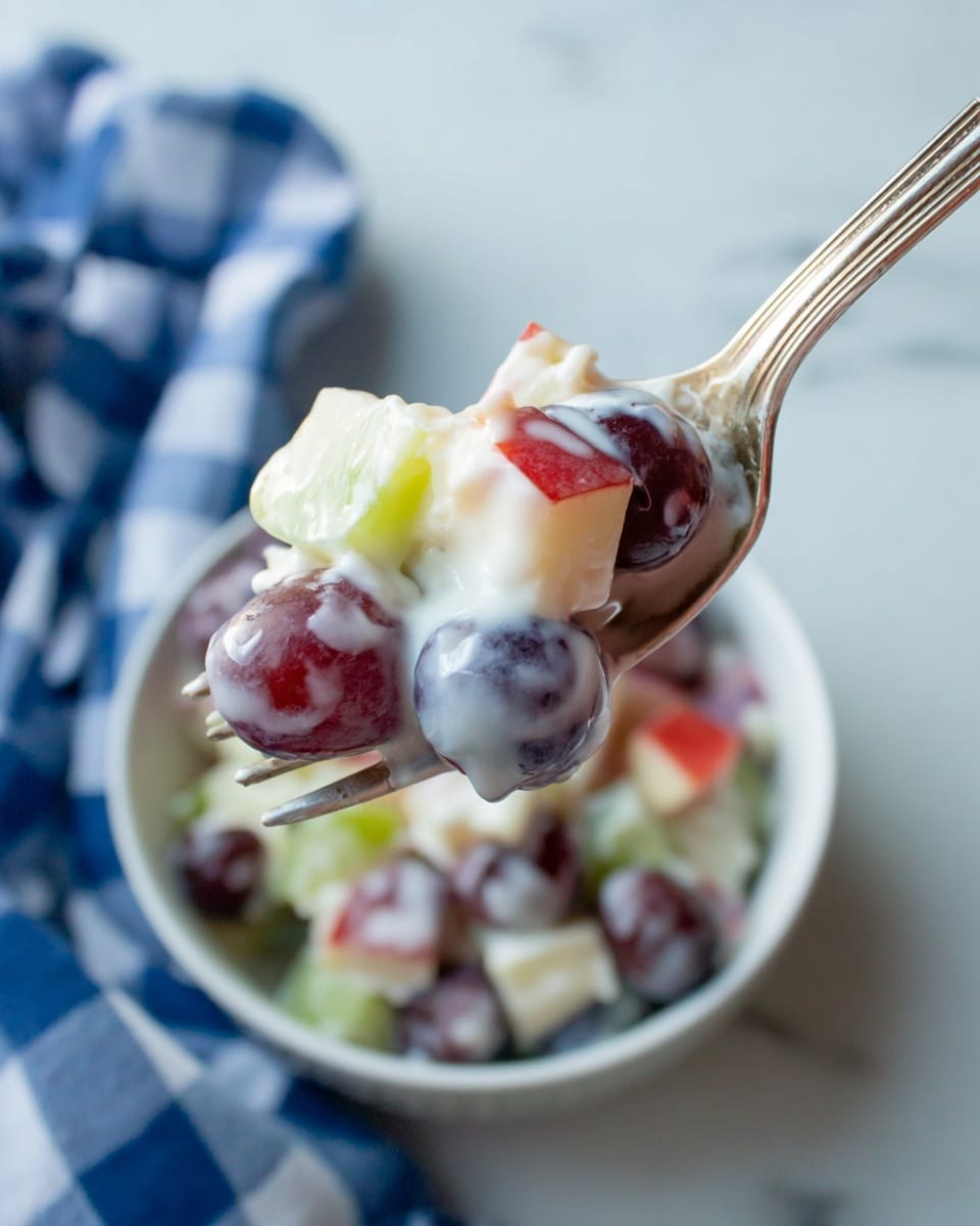 A close-up view of a silver fork holding a mix of small fruit pieces covered in creamy yogurt. The fruit pieces include purple grapes, green apple chunks, and red apple bits, all coated with white yogurt showing a shiny, smooth texture. In the background, there is a white bowl filled with the same fruit and yogurt mix. The scene includes a crumpled blue and white checkered cloth beside the bowl on a white marbled surface. photo taken with an iphone --ar 4:5 --v 7