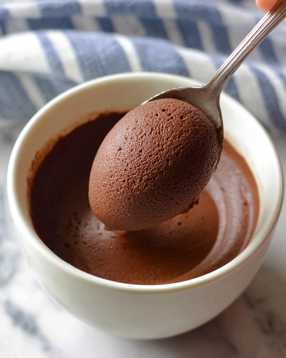 The image shows a close view of a white bowl filled with smooth, dark brown chocolate mousse. The mousse has a soft, creamy texture and fills the bowl almost to the top. A spoon is lifting a rounded scoop of the mousse, showing its light and airy texture. The background features a white marbled surface with a blue and white striped cloth partially visible. A woman's hand is holding the spoon at the bottom of the frame. photo taken with an iphone --ar 4:5 --v 7