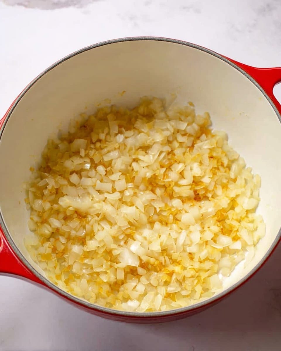 Inside a white pot with a red outer side, there is one layer of small chopped onions that are light golden brown and soft. The onions cover the bottom of the pot evenly, with some parts slightly more cooked showing a deeper yellow color. The texture looks tender and slightly translucent. The inside of the pot is white, and the surface below the pot is a white marbled texture. photo taken with an iphone --ar 4:5 --v 7