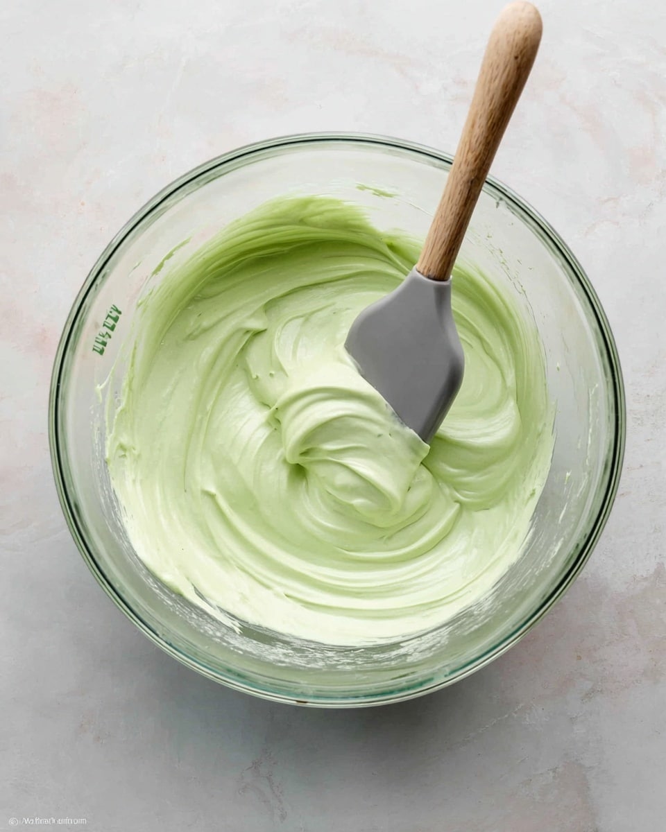 A clear glass bowl is placed on a white marbled surface, filled with a thick, smooth light green creamy mixture taking up one layer. A spatula with a wooden handle and a gray silicone head is partially dipped into the cream on the right side of the bowl, creating soft swirls and texture on the surface of the mixture. The bowl is seen from directly above, showing its round shape and clear glass handle on the left side. Photo taken with an iphone --ar 4:5 --v 7
