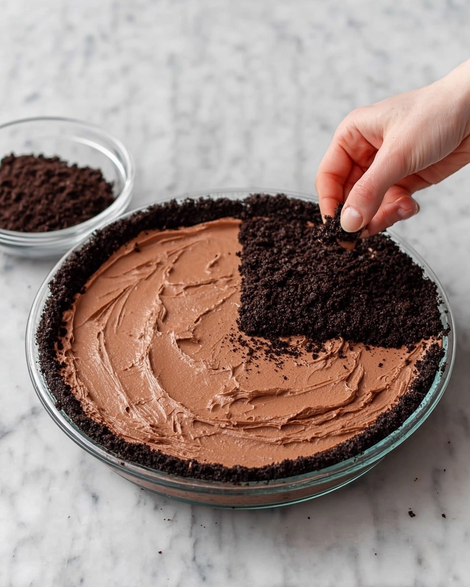 A round pie dish with a dark crumb crust forms the base and sides, filled with a smooth, thick layer of light brown chocolate filling spread evenly. On the right side, a woman's hand sprinkles a generous layer of dark crumb topping over the chocolate filling from a small clear bowl beside the dish, partially covering the filling. The surface is a white marbled texture. Photo taken with an iphone --ar 4:5 --v 7