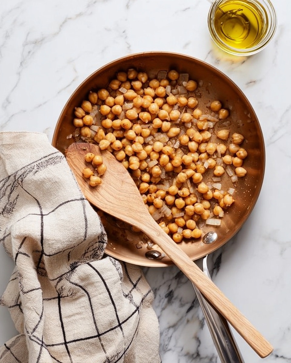 A round metal pan with a golden-brown color holds a single layer of cooked chickpeas mixed with translucent diced onions. Resting diagonally on top of the chickpeas is a smooth, light wooden spoon with a round end. Under the pan’s handle is a crumpled beige cloth with black grid lines. To the top right of the pan on a white marbled surface sits a small glass jar filled with golden olive oil. photo taken with an iphone --ar 4:5 --v 7