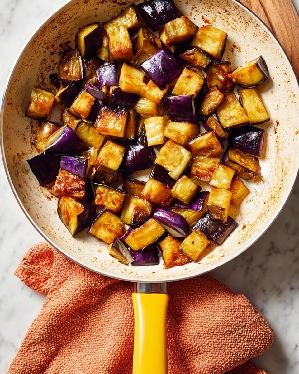 The image shows a white pan filled with golden brown and deep purple chunks of cooked eggplant, lightly browned from frying. The eggplant pieces are uneven in size and scattered across the pan, showing slightly crispy edges and soft, tender centers with a shiny, oily texture. The pan has a yellow handle resting on an orange textured cloth, all placed on a white marbled surface. The lighting highlights the rich, warm tones of the eggplant and the pan's worn cooking marks, creating a homey and inviting feel. photo taken with an iphone --ar 4:5 --v 7