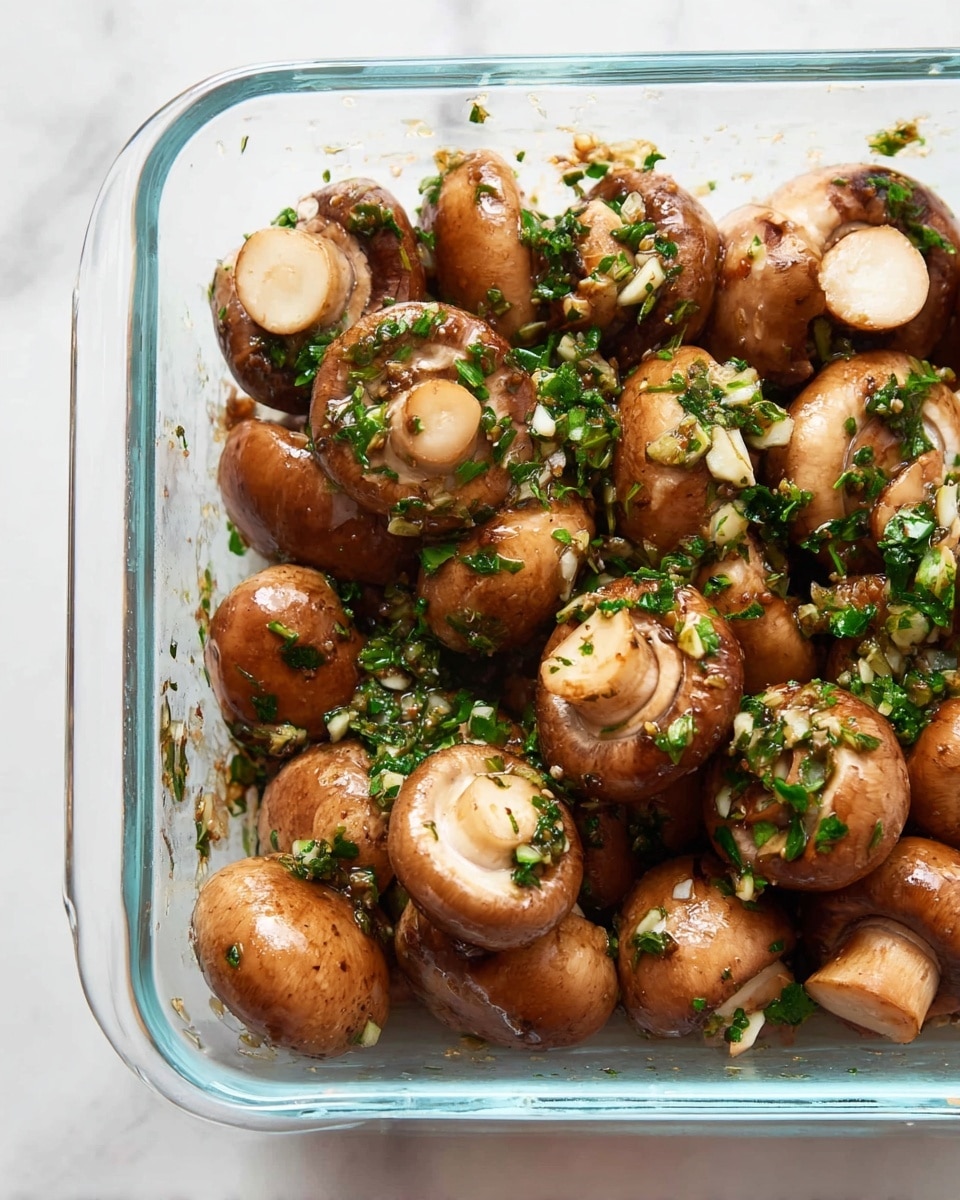 The image shows a clear glass rectangular dish filled with medium-sized brown mushrooms, whole and halved, with their smooth caps and white stems visible. The mushrooms are coated with bits of bright green herbs and garlic pieces scattered mostly on top. The dish sits on a white marbled surface. The mushrooms have a shiny, slightly wet texture indicating they are seasoned or marinated. photo taken with an iphone --ar 4:5 --v 7