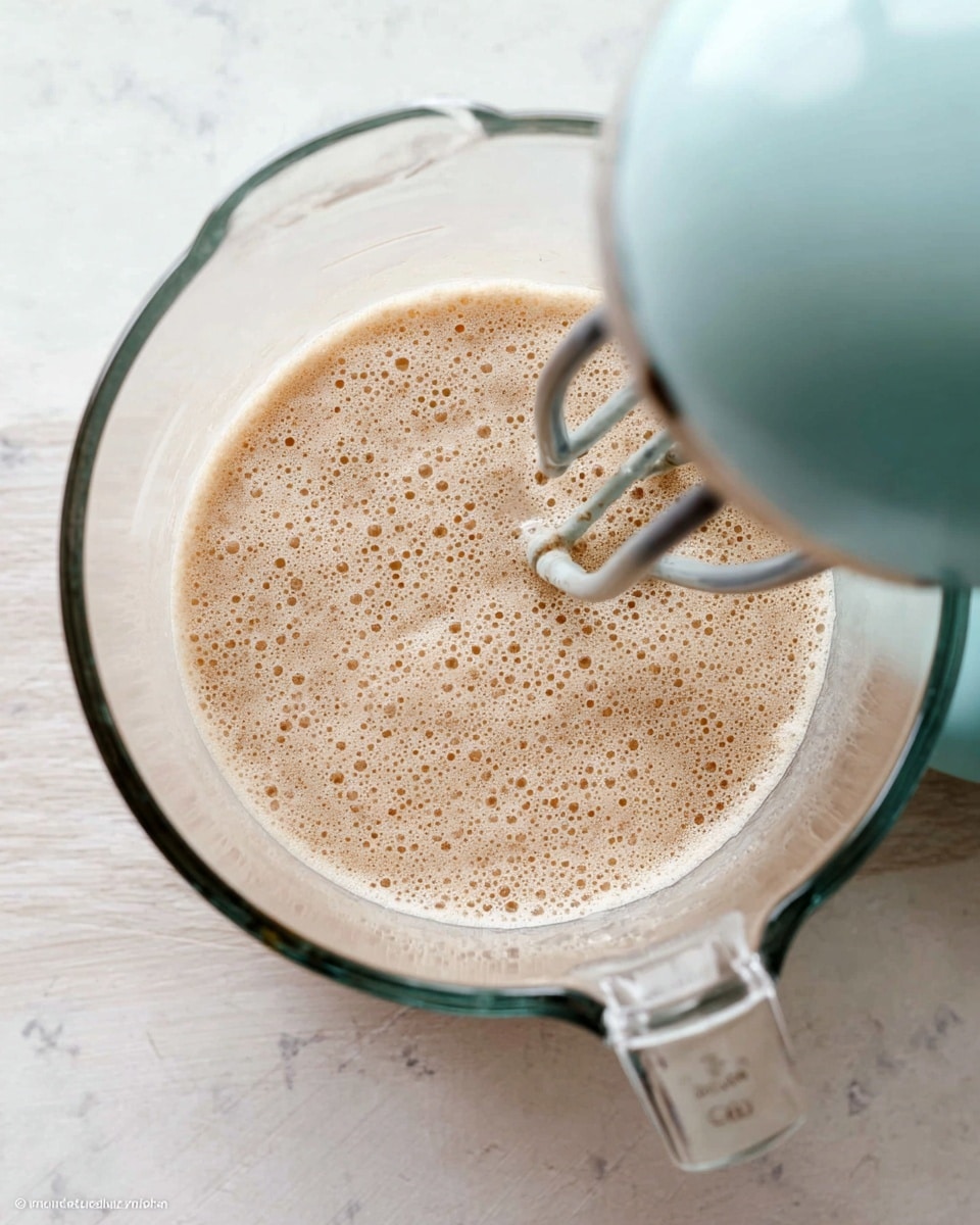 A clear glass mixing bowl holds a light beige frothy mixture with many bubbles on top, filling the bottom layer of the bowl. Over the bowl, a light blue electric mixer is positioned on the right side with one black beater attachment partially visible inside the bowl. The bowl rests on a white marbled surface. photo taken with an iphone --ar 4:5 --v 7