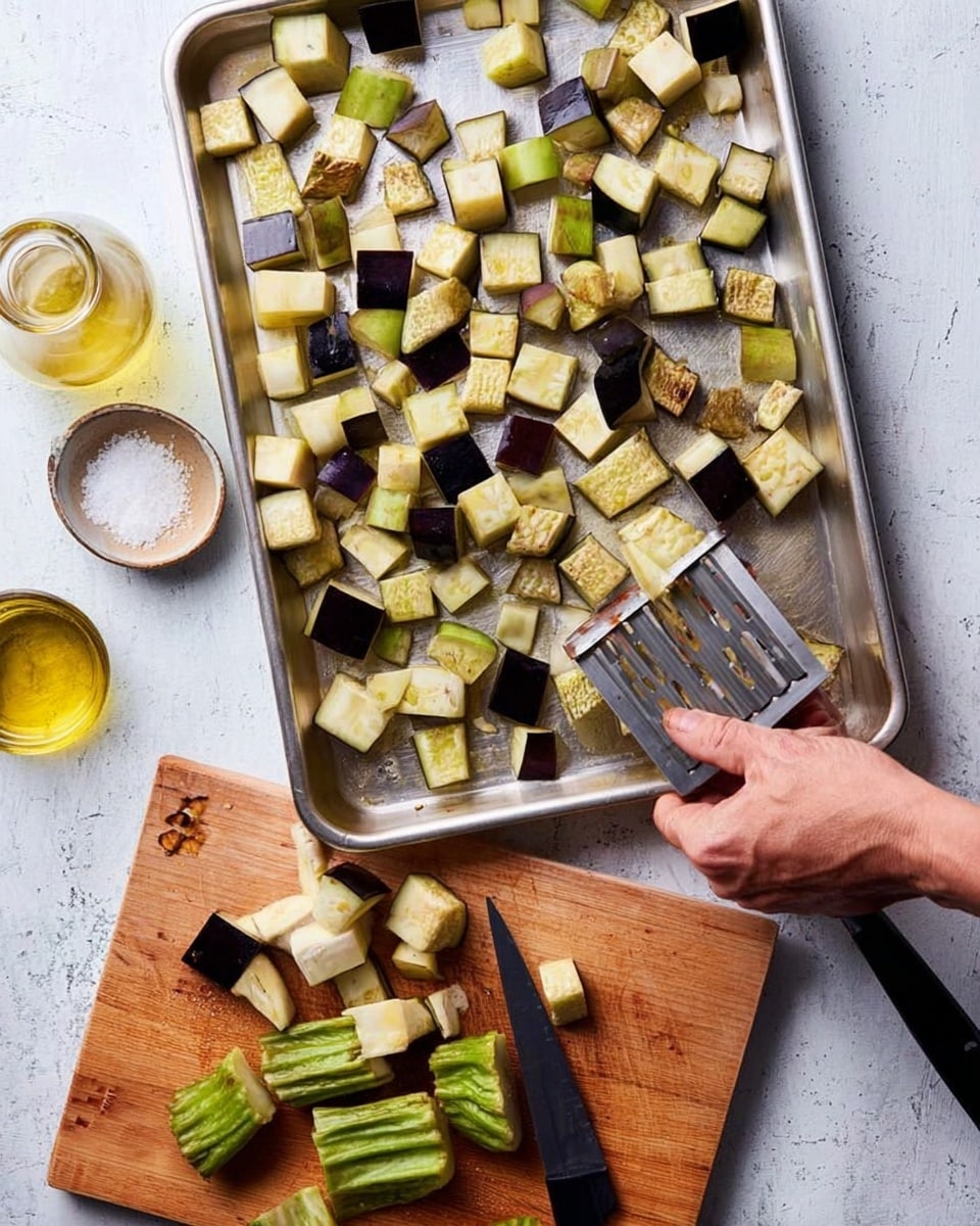 A metal tray filled with cubed eggplant pieces in light green and dark purple shades, some showing browned edges, sits on a white marbled surface. A woman's hand holding a silver potato masher is pressing down on the eggplant cubes. Below the tray, a wooden cutting board holds the green, wrinkled eggplant top, a black-handled knife, and more cubed eggplant pieces. Next to the board is a small bowl of coarse salt, and to the left of the tray is a clear glass bottle filled with golden olive oil. Photo taken with an iphone --ar 4:5 --v 7
