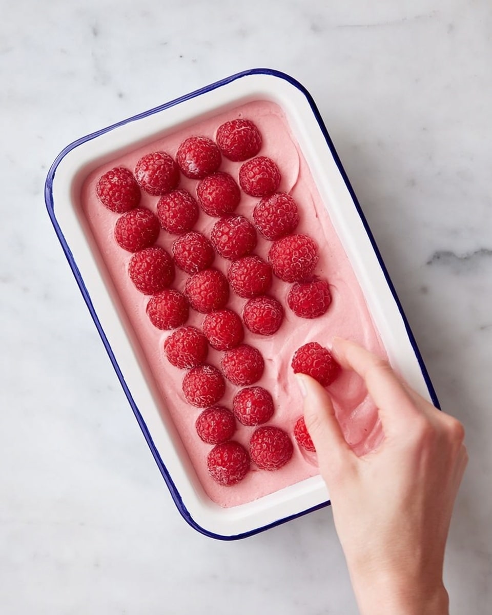 A white rectangular dish with a blue rim is filled with a smooth pink creamy layer spread evenly inside. On top of this pink layer, there are two rows of fresh red raspberries neatly placed in a grid pattern, covering most of the surface. A woman's hand is gently placing another raspberry near the bottom right corner of the dish. The dish is set on a white marbled surface, adding a clean and fresh look to the scene. photo taken with an iphone --ar 4:5 --v 7