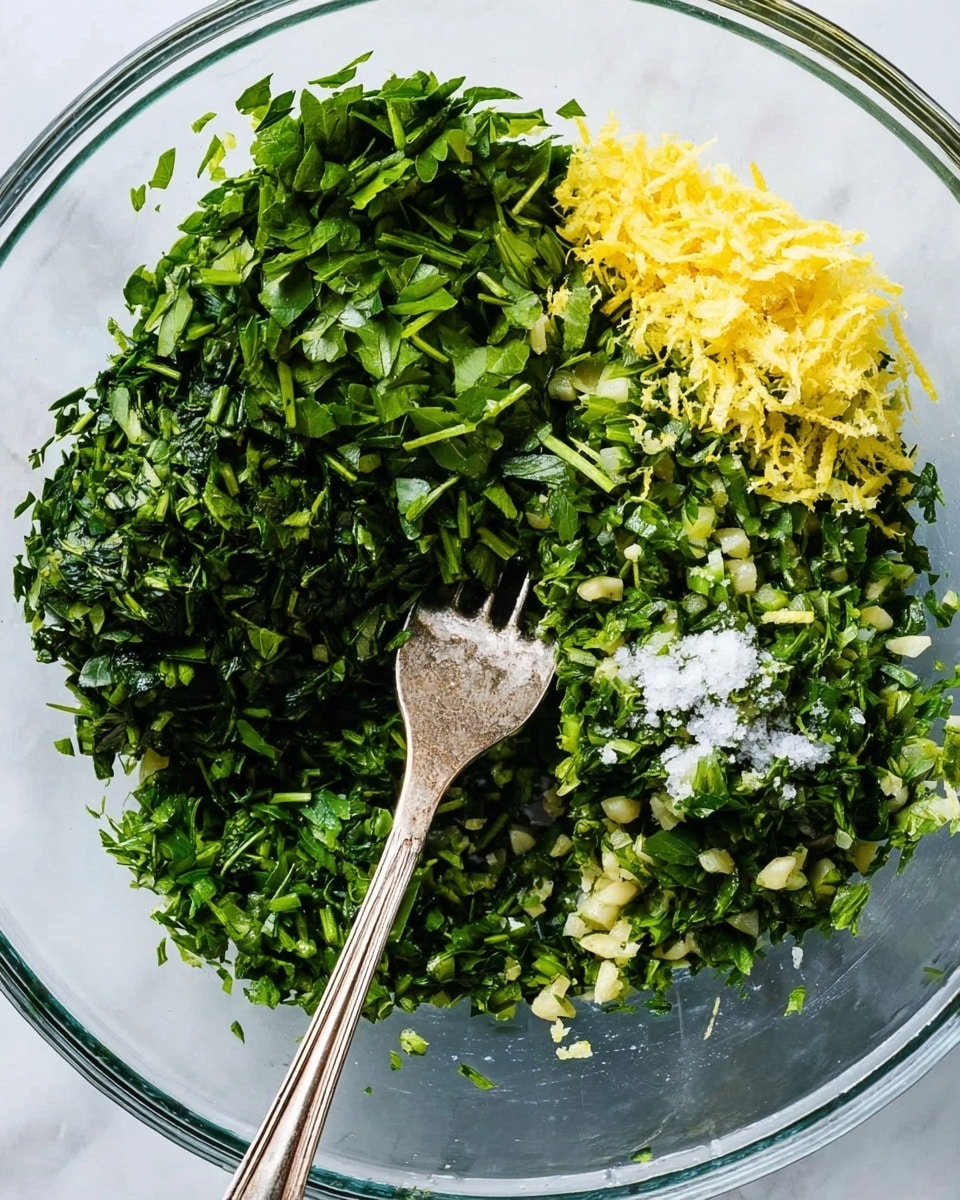 A close-up top view of a clear bowl with four main layers of ingredients separated but close together. The largest layer is finely chopped dark green herbs filling about two-thirds of the bowl, with bright green chopped parsley. To the right, there is a small pile of yellow grated lemon zest and a few bits of white coarse salt on top of chopped herbs. Below, there is a small mound of pale minced garlic on more chopped dark green herbs. To the left, there is another pile of finely chopped dark green herbs. A silver fork rests diagonally over the herbs in the bowl. The bowl is on a white marbled surface. Photo taken with an iphone --ar 4:5 --v 7