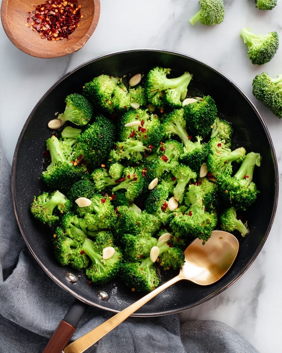 The image shows a shallow black pan filled with bright green broccoli florets scattered evenly across the surface. Thin slices of light beige garlic are spread on top of the broccoli, along with small bits of red chili flakes providing a touch of color contrast. A shiny golden spoon lies on the right side of the pan, partially resting on the broccoli. The pan is placed on a white marbled surface, with a folded grey cloth visible on the left side. In the upper left corner, part of a small wooden dish with red chili flakes is visible, and in the upper right corner, a few broccoli florets rest on the white marbled surface. Photo taken with an iphone --ar 4:5 --v 7