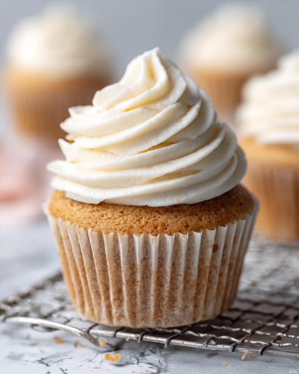 The image shows a close-up of a single cupcake placed on a metal wire rack. The cupcake has a light brown base with a soft texture, wrapped in a slightly translucent paper liner. On top, there is a thick swirl of white cream frosting with soft peaks, smoothly piped in a spiral shape. The background is a white marbled surface with a blurry, soft focus on more cupcakes in the distance. photo taken with an iphone --ar 4:5 --v 7