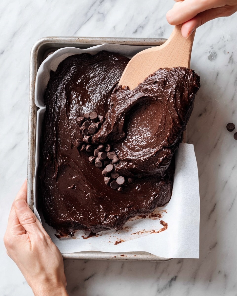 A close-up of thick, dark brown chocolate batter with small chocolate chips scattered inside, being spread on white parchment paper in a metal tray. The texture looks smooth and glossy with some rough spots from the chips. Two woman's hands are visible, one holding the tray and the other using a wooden spatula to spread or lift the batter. The background features a white marbled surface. photo taken with an iphone --ar 4:5 --v 7