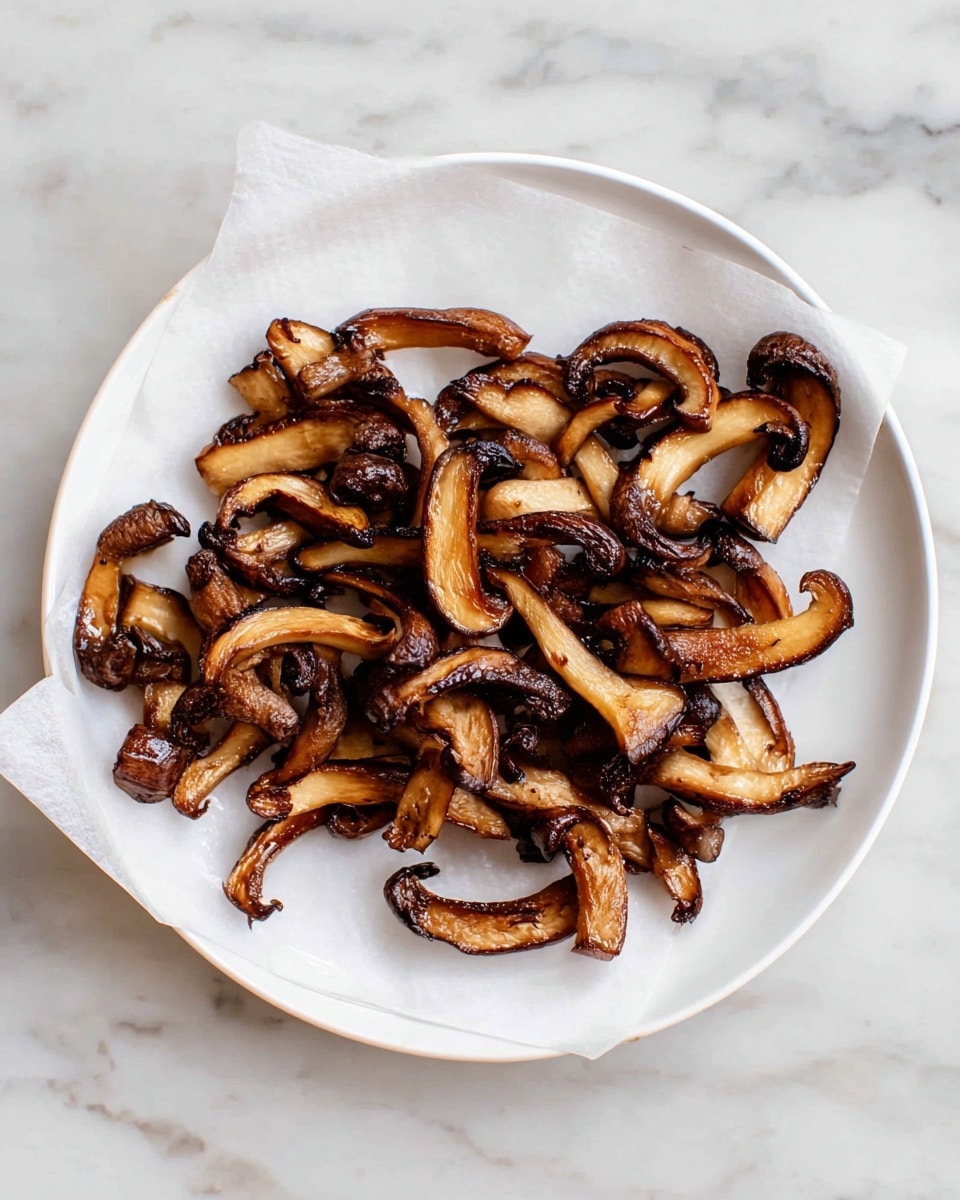 A white plate holds a single layer of browned, sliced mushrooms with dark edges and a slightly shiny texture, some curved and others more flat, sitting on a sheet of white parchment paper. The plate is placed on a white marbled surface, showing light grey and subtle veining throughout. The mushrooms are cooked and have a rich brown color with hints of caramelization, creating a contrast against the white paper and plate. photo taken with an iphone --ar 4:5 --v 7
