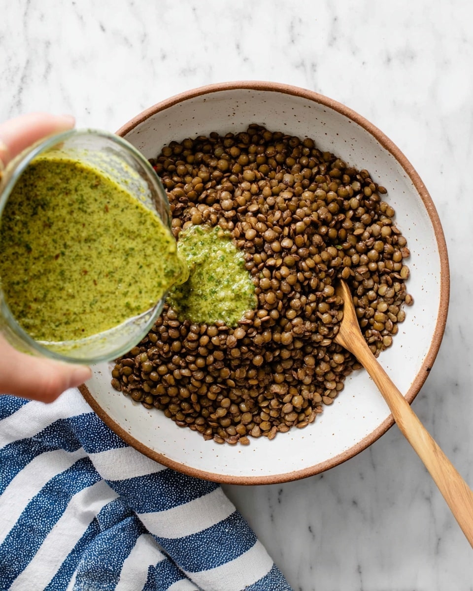 A white bowl with a brown rim holds a single thick layer of cooked brown lentils, textured and piled mostly to the right side. A wooden spoon rests inside the bowl on the right, partially covered by the lentils. On the left, a woman's hand is pouring a bright green, slightly chunky sauce from a clear measuring cup into the bowl. The scene is set on a white marbled textured surface with a blue and white striped cloth underneath the bowl. photo taken with an iphone --ar 4:5 --v 7