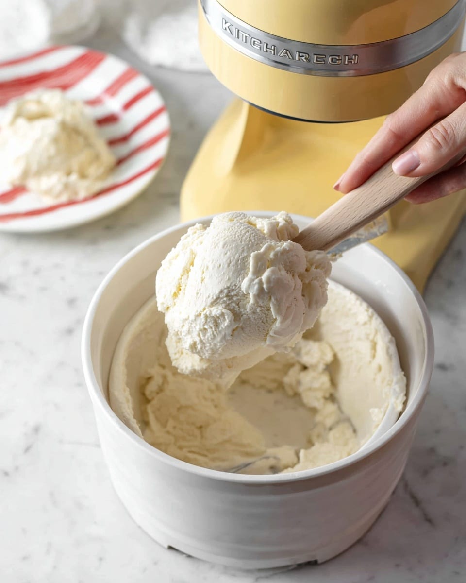 A close-up of a yellow KitchenAid ice cream maker with thick, creamy white ice cream inside the white bowl. A woman's hand holds a wooden spatula lifting a large scoop of the textured ice cream from the machine. In the background, a white plate with red stripes holds more of the creamy ice cream. The whole scene is set on a white marbled surface. photo taken with an iphone --ar 4:5 --v 7