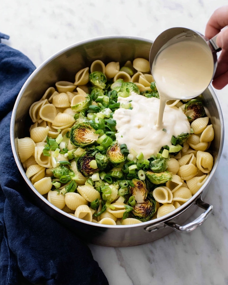 A silver pot sits on a white marbled surface with a dark blue cloth beside it. Inside the pot, there are small shell-shaped pasta pieces, bright green chopped scallions, and halved roasted Brussels sprouts with a slightly charred texture. A creamy white sauce is being poured from a small metal measuring cup into the center of the pot, covering part of the pasta and vegetables. A woman's hand holds the measuring cup from the right side of the image. photo taken with an iphone --ar 4:5 --v 7