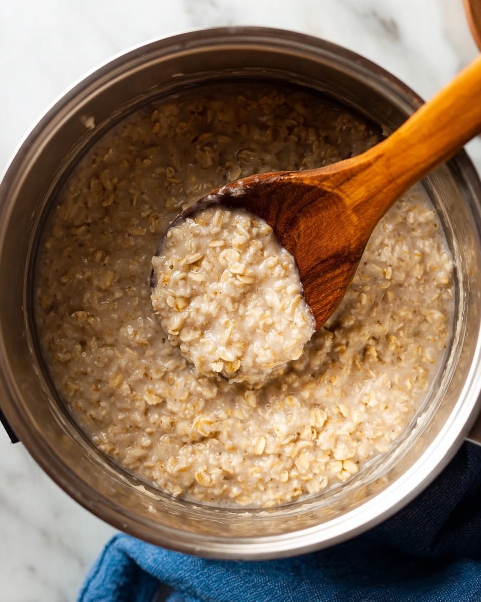 A close-up view inside a metal pot filled with thick cooked oatmeal that has a light brown and creamy texture, with small visible oat grains spread throughout the mixture. A wooden spoon with a rich brown color is placed inside the pot, slightly lifting some oatmeal, showing the creamy and soft texture clearly. The pot sits on a white marbled surface with a blue cloth partially visible at the bottom. The clear metal sides of the pot reflect light softly with a smooth finish. Photo taken with an iphone --ar 4:5 --v 7