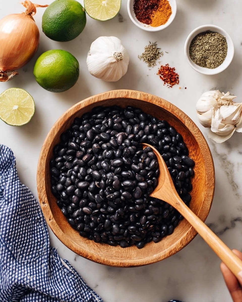 A wooden bowl filled with shiny black beans takes up the middle of the image, with a wooden spoon resting inside it. Around the bowl on a white marbled surface are fresh limes, a white onion, a garlic bulb, and small white bowls filled with different spices, one with green dried herbs and another with red and brown spice powders. A woman's hand is not visible, but a blue and white checkered cloth is draped on the left side of the frame. Photo taken with an iphone --ar 4:5 --v 7