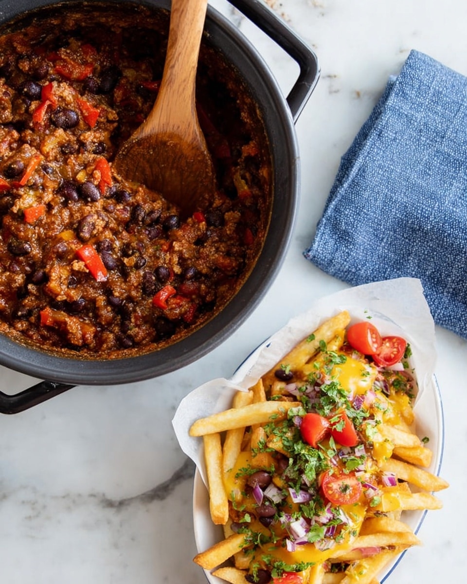 A black pot with two handles is filled with a thick chili mix that has visible red bell pepper pieces, black beans, and a rich brown sauce. A wooden spoon rests inside the pot. Below, a white plate lined with white parchment paper holds a serving of fries covered in melted yellow cheese sauce, chili, halved red cherry tomatoes, small green chili slices, chopped red onions, and green herbs. The setup is on a white marbled surface with a blue cloth beside the pot and some green herb sprigs around. Photo taken with an iphone --ar 4:5 --v 7